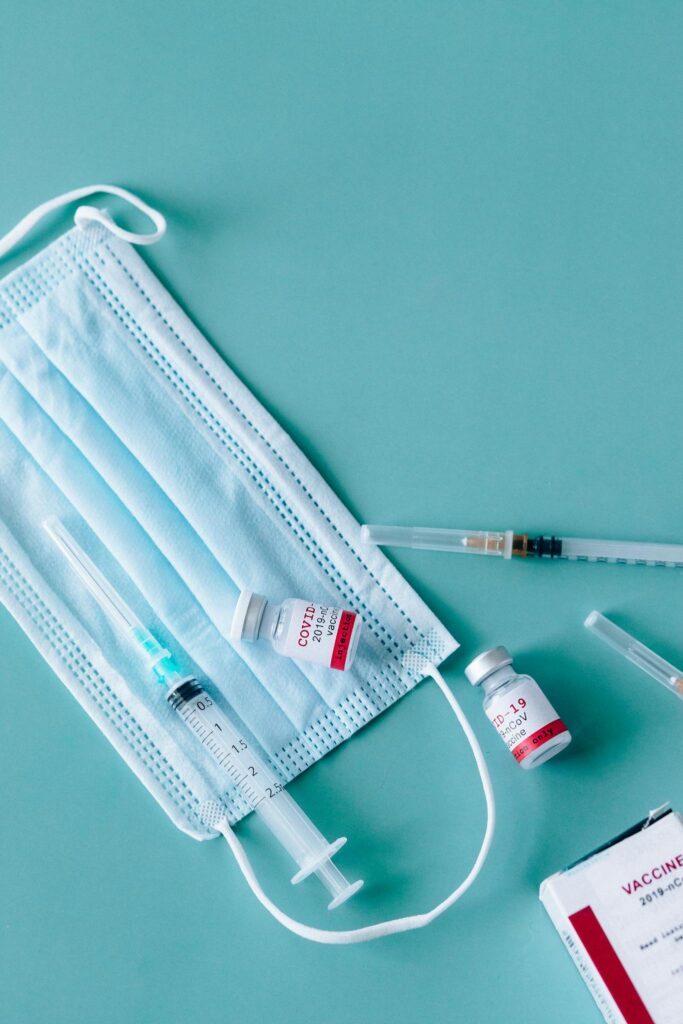 Medical supplies including face masks, syringes, and COVID-19 vaccine vials on a sterile teal background.